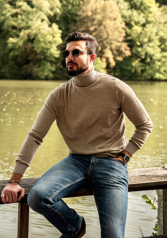 homme avec col roulé beige avec un jeans assis sur une poutre le long d'une rivière